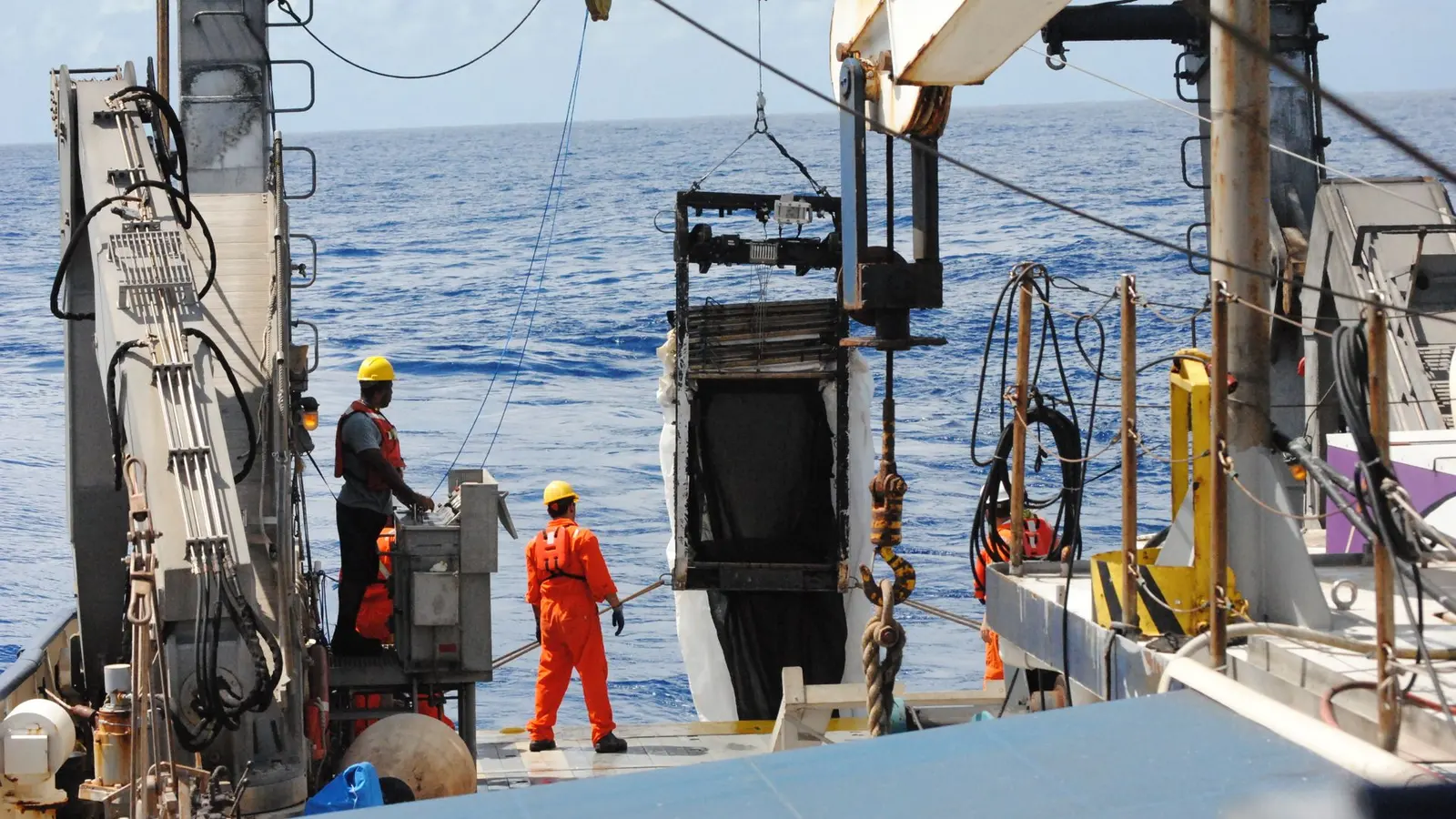 Two crew members in hard hats and life vests lowering equipment from a vessel deck into open sea